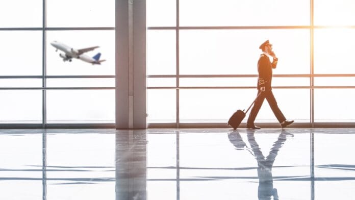 A uniformed pilot walking through an airport terminal while talking on a phone, with a departing aircraft visible through the floor-to-ceiling windows behind them.