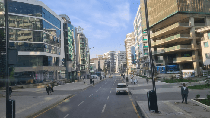 Wide street view of central Addis Ababa lined with modern glass-fronted buildings, a partly constructed high-rise, a few pedestrians, and light traffic under a blue sky.